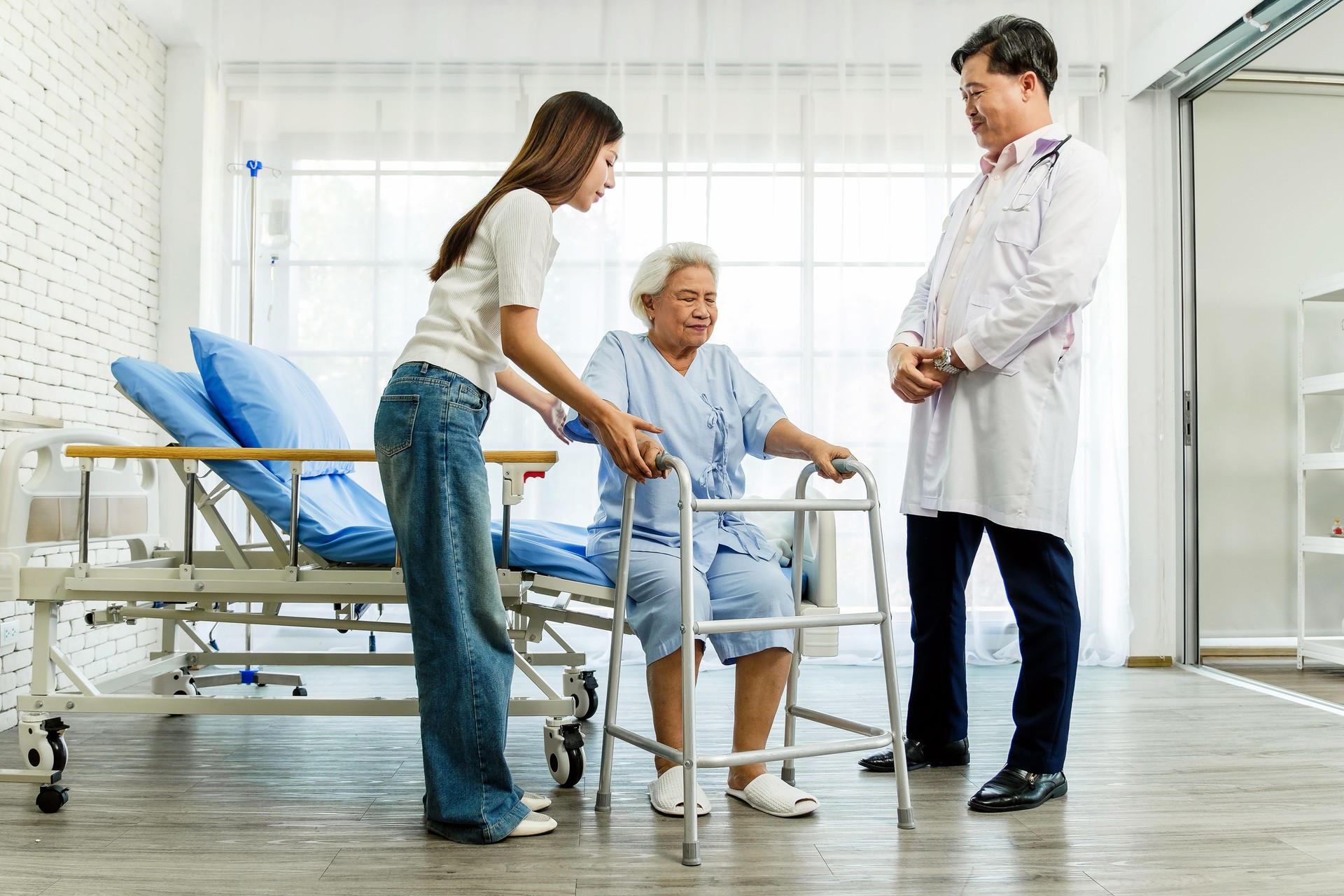 Senior woman using a walker with help from a caregiver and support from a doctor in a clinic. Represents rehabilitation, elderly care, mobility support, and recovery in a medical setting.